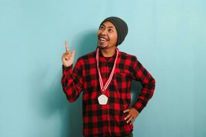 trots jong Aziatisch Mens points omhoog terwijl Holding een medaille, vieren, geïsoleerd Aan blauw achtergrond. foto