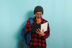 een attent jong Aziatisch leerling Mens met muts en rood plaid flanel shirt, vervelend een rugzak en bril, is denken terwijl Holding boeken op zoek voor ideeën terwijl staand tegen een blauw achtergrond foto