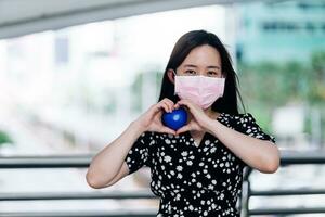 Aziatisch vrouw vervelend masker en Holding een blauw hart in de stad. foto