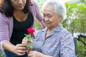 Aziatisch ouderen vrouw Holding rood roos bloem, glimlach en gelukkig in de zonnig tuin. foto
