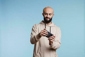 jong Arabisch Mens spelen mobiel spel, gebruik makend van onderhoudend toepassing software. geconcentreerd gamer genieten van videogame Aan smartphone terwijl staand in studio Aan blauw achtergrond foto