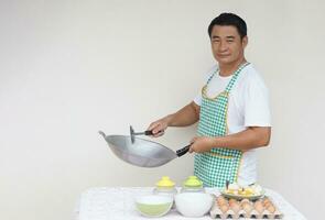 knap Aziatisch Mens chef is Koken gebakken eieren, draagt wit overhemd en schort, houdt frituren pan en pollepel spatel . concept, liefde Koken. keuken levensstijl. foto