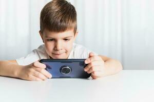 een weinig jongen in een wit t-shirt Toneelstukken spellen Aan de telefoon Bij huis. een gelukkig kind looks Bij zijn smartphone. foto