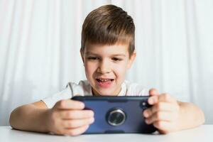 een weinig jongen in een wit t-shirt Toneelstukken spellen Aan de telefoon Bij huis. een gelukkig kind looks Bij zijn smartphone. foto