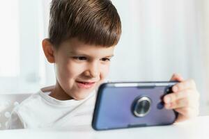 een weinig jongen in een wit t-shirt Toneelstukken spellen Aan de telefoon Bij huis. een gelukkig kind looks Bij zijn smartphone. foto