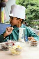 schattig Indisch chef jongen voorbereidingen treffen ijscoupe schotel net zo een een deel van niet brand Koken welke omvat vanille ijs room, brownie, kokosnoot poeder, vers gehakt fruit en aardbei siroop. weinig kind voorbereidingen treffen voedsel foto