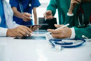 medisch team hebben een vergadering met artsen in wit laboratorium jassen en chirurgisch scrubs gezeten Bij een tafel bespreken een patiënten werken online gebruik makend van computers foto