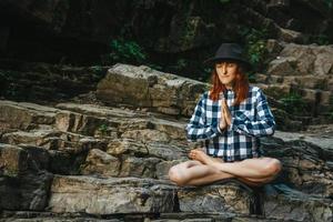 vrouw in een hoed en shirt mediterend op rotsen in een lotushouding foto
