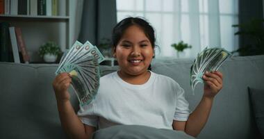 portret van vrolijk tiener meisje tonen dollar Bij camera en glimlachen in de leven kamer Bij huis. foto