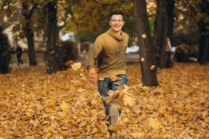 knappe kerel lacht en rent gele bladeren in het herfstpark verstrooid foto