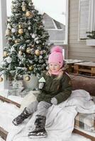 een weinig meisje in winter kleren is zittend Aan de veranda van een huis versierd voor de Kerstmis vakantie. Kerstmis vakantie voor een kind. foto
