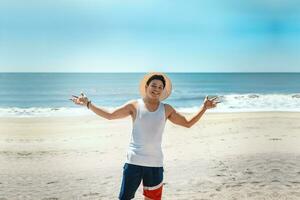 concept van Mens genieten van de strand. portret van Mens in hoed Aan de strand verspreiden zijn armen. glimlachen jong Mens Aan de strand verspreiden armen op zoek Bij camera foto