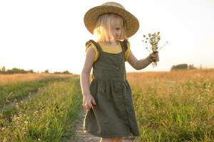 een weinig blond meisje in een rietje hoed wandelingen in een veld- met een boeket van madeliefjes. foto