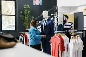 elegant vrouw boodschappen doen voor formeel dragen, op zoek Bij modieus pak in kleding op te slaan. Afrikaanse Amerikaans klant buying modieus handelswaar, analyseren kleren kleding stof in modern winkel foto