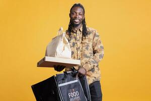 portret van restaurant arbeider Holding papier zak leveren meenemen voedsel naar klanten gedurende lunch tijd. Afrikaanse Amerikaans postbode draag- thermisch rugzak vol met snel voedsel in studio foto