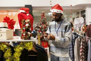 op te slaan assistent vervelend de kerstman hoed regelen van kleren Aan rekken voor seizoensgebonden aanbiedingen en promoties, Kerstmis versierd kleding op te slaan. leidinggevende op zoek Bij accessoires in kleinhandel winkel gedurende Kerstmis. foto