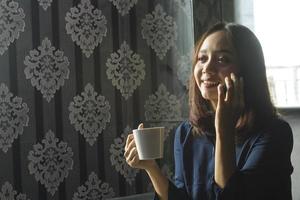 lachende aziatische vrouw die koffie drinkt terwijl ze een mobiele telefoon gebruikt voor werk foto