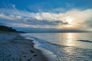 zonsondergang Aan de west strand Aan de Baltisch zee. golven, strand, bewolkt lucht en zonneschijn foto