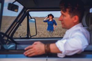 man rijdende auto en vrouw in jurk en hoed wandelen door tarweveld