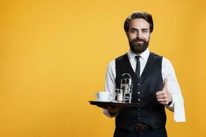 gelukkig ober doet duimen omhoog teken terwijl hij houdt schotel met koffie en accessoires in studio, professioneel elegant butler. Mens restaurant werknemer tonen Leuk vinden overeenkomst gebaar Aan camera. foto