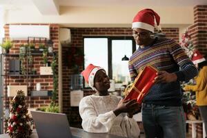 bedrijf werknemer geven Kerstmis geschenk naar collega en tonen naam kaart gehecht naar doos. Afrikaanse Amerikaans Mens en vrouw collega's uitwisselen Kerstmis presenteert in feestelijk versierd kantoor foto