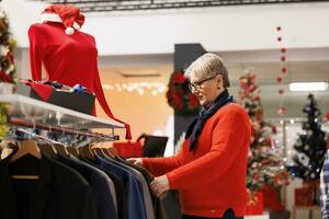 senior cliënt zoeken voor kleren in mode op te slaan gedurende seizoensgebonden promoties, op zoek voor Kerstmis avondeten kleding in winkel met feestelijk decor. vrouw klant Bij boodschappen doen centrum koopt geschenken. foto