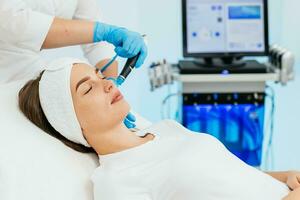 schoonheidsspecialiste kliniek. professioneel vrouw schoonheidsspecialist aan het doen hydrafaciale procedure terwijl wezen een werk. aantrekkelijk mooi hoor vrouw aan het liegen Aan de medisch bed terwijl hebben schoonheid procedures foto