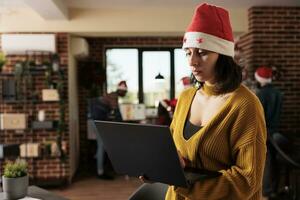 vrouw project manager vervelend de kerstman hoed beheren taken Aan laptop terwijl staand in winter feestelijk kantoor. werknemer Holding portable computer en werken voor deadline Bij Kerstmis vooravond foto