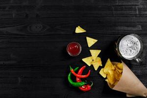 top visie van tortilla chips met saus, glas van bier en rood Chili peper Aan zwart houten achtergrond foto