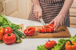 jong vrouw Koken in de keuken Bij huis. een vrouw bezuinigingen een peper en groenten met een mes. foto