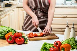 jong vrouw Koken in de keuken Bij huis. een vrouw bezuinigingen een tomaat en groenten met een mes. foto