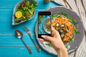 fotograferen voedsel concept - vrouw duurt afbeelding van pasta met aubergines, tomaat, kaas, rucola en salade foto