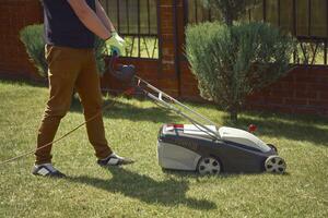 Mens in gewoontjes kleren en handschoenen is gras maaien gras met modern gazon maaier Aan zijn achtertuin. tuinieren zorg uitrusting en Diensten. zonnig dag foto