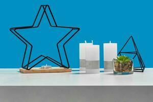 wit tafel met groen sappig, drie groot kaarsen en een klein in kandelaar in het formulier van ijzer ster, zwart driehoek. blauw achtergrond. dichtbij omhoog foto