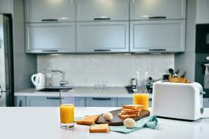 tosti apparaat met vers geroosterd brood, eieren en glas van oranje sap Aan een licht keuken tafel foto