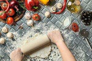 vrouw handen rollen uit deeg Aan metaal achtergrond, dichtbij omhoog. chef maakt deeg. tafel met groenten en keukengerei. Italiaans keuken concept. pizza foto