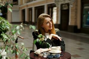 mooi blond vrouw in elegant gewoontjes bedrijf kleding zittend in cafe in mooi Europese stad. gebruik makend van mobiel telefoon. foto