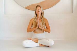 binnen- foto van jong blond vrouw aan het doen yoga opdrachten en beoefenen meditatie. zittend in lotos houding in yoga klas.
