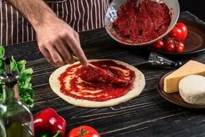 een mannetje hand- verspreiden tomaat puree Aan een pizza baseren met lepel Aan een oud houten achtergrond foto