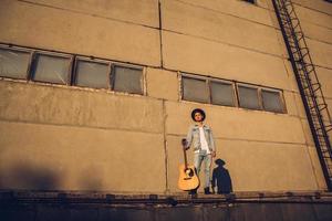 jonge kerel muzikant in een hoed met een gitaar tegen een muur van beton foto