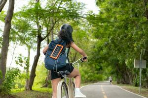 achterzijde visie van Aziatisch vrouw rijden een fiets in de park. het heeft een rugzak met een zonne- paneel voor opladen smartphone. foto