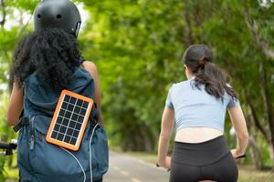 beide van jong Dames in sportkleding rijden een fiets in de park, dragen een rugzak met een zonne- paneel naar in rekening brengen smartphone. foto