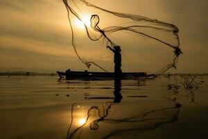 silhouet van visser Bij zonsopkomst, staand aan boord een roeien boot en gieten een netto naar vangst vis voor voedsel foto