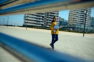 zelfverzekerd jong vrouw ondernemer in gewoontjes kleding, Holding laptop, glimlachen op zoek Bij camera, wandelen de straat foto