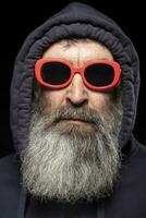 een ouderen Mens met een grijs baard in een kap en rood bril Aan een zwart achtergrond. een Mens verbergt iets. foto