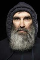 een ouderen Mens met een grijs baard in een kap Aan een zwart achtergrond. foto