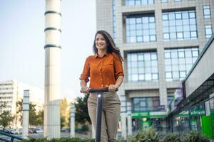 De volgende generatie pendelen naar werk gebruik makend van e-scooters door vrouw bedrijf personen. jong mooi vrouw in een blauw jasje glimlacht en ritten een elektrisch scooter naar werk langs kantoor gebouwen foto