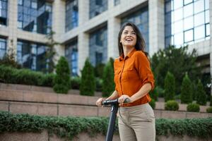 jong mooi vrouw glimlacht en ritten een elektrisch scooter naar werk langs kantoor gebouwen. ecologisch vervoer concept. foto