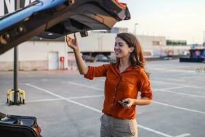 jong mooi modieus vrouw bestuurder is opening auto romp glimlachen Bij parkeren kavel, vrouw sluitend auto romp geparkeerd staand door de terug romp van haar auto Aan de parkeren veel foto