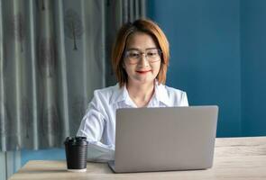 Aziatisch vrouw zittend Aan een stoel terwijl gebruik makend van laptop werken en typen een e-mail Aan laptop Bij huis. foto